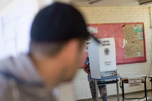 Um homem de costas e de boné preto, borrado pela lente da câmara, olha para outro, dentro de uma sala de votação, que está votando detrás da cabine de votação.