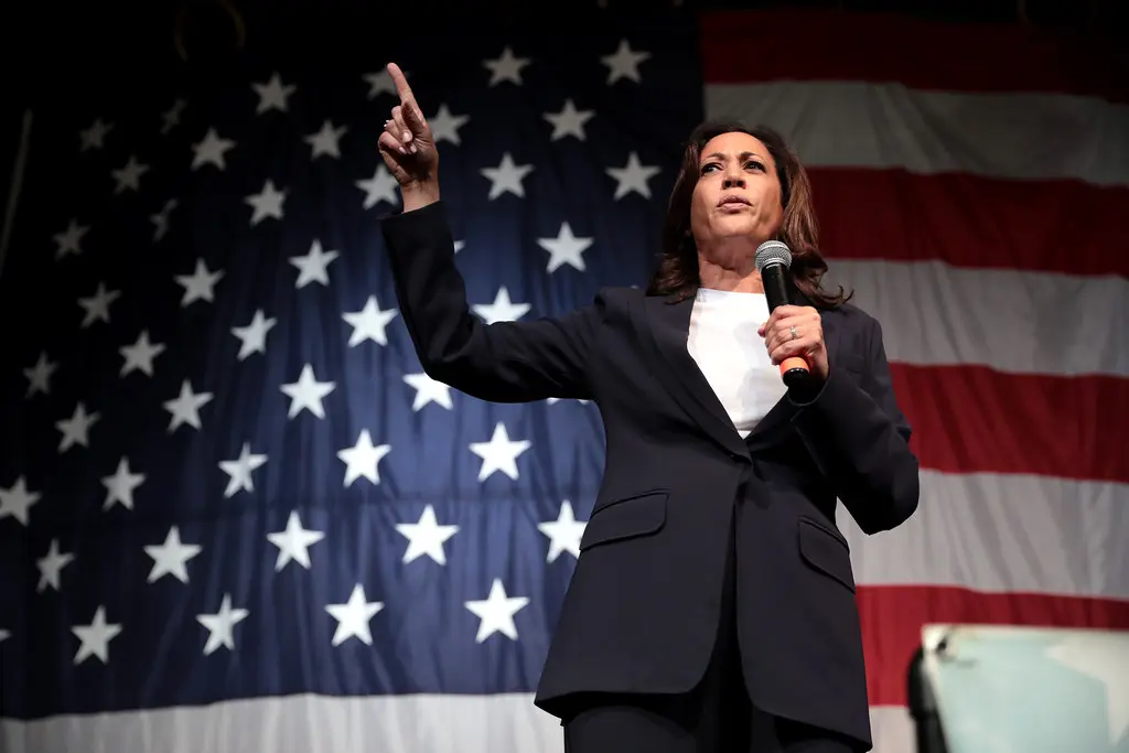 Kamala Harris discursa durante um comício. Com a mão direita, aponta para frente, enquanto segura um microfone com a esquerda. Ao fundo, a bandeira dos Estados Unidos.