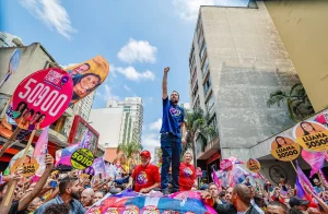 O candidato Guilherme Boulos com seu punho direito erguido, em cima de um caminhão de som, no centro de São Paulo. Ao seu redor, dezenas de apoiadores, sua vice, Marta Suplicy, e o presidente Lula.